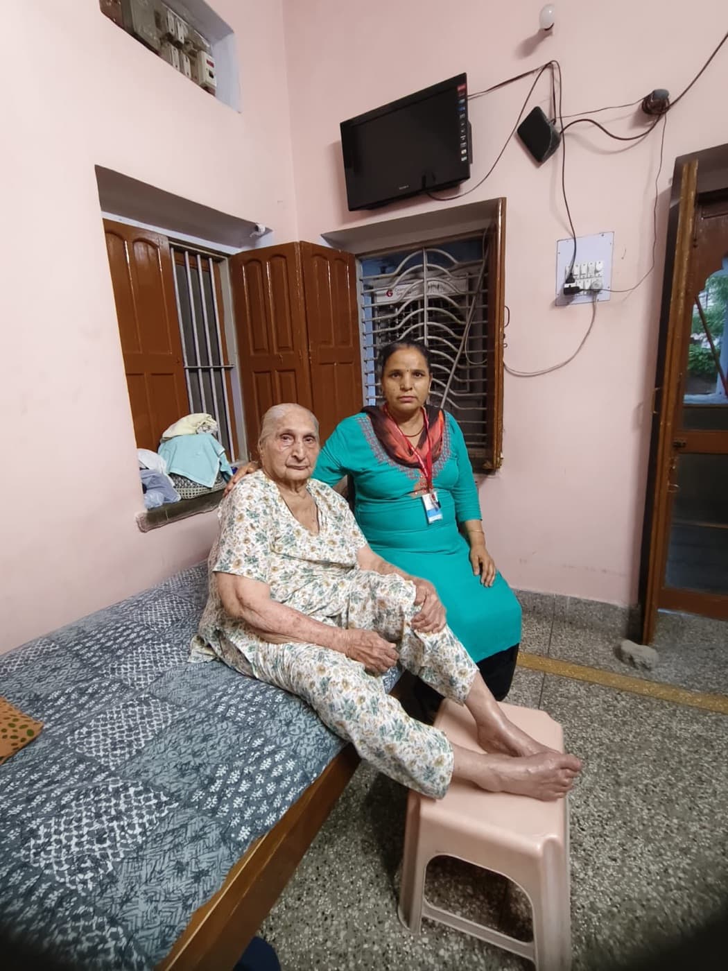 An Advino caregiver sitting with an elderly woman on the edge of her bed at home.