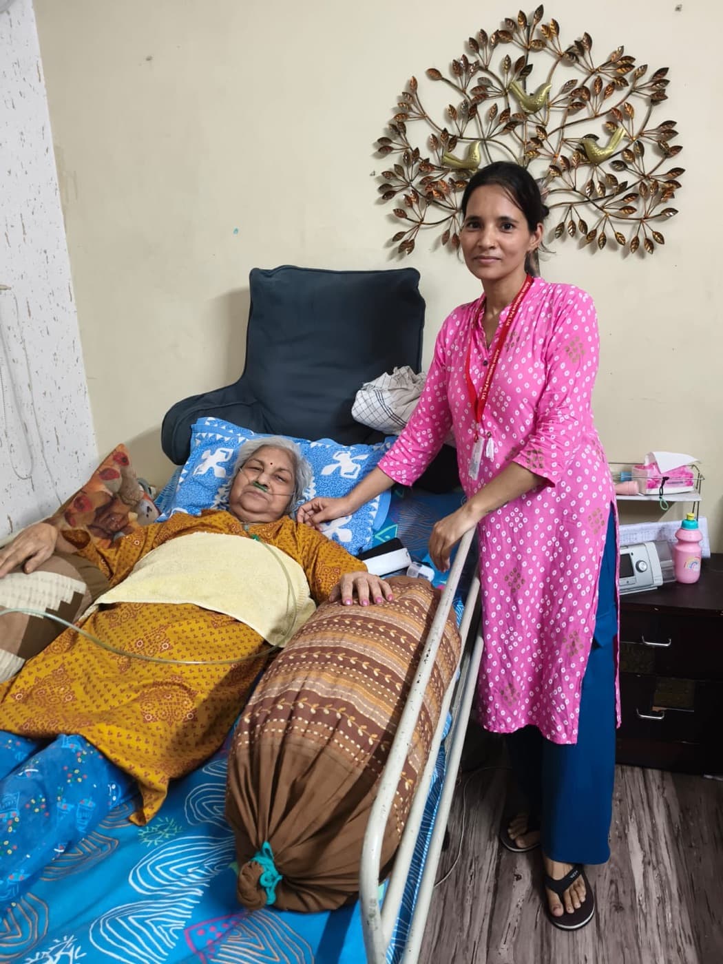 An Advino caregiver standing at the bedside of an elderly woman being cared for at home.